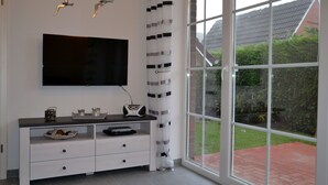 A modern living room with a flat-screen TV mounted on the wall, a white entertainment unit with drawers, and a large window with a view of a brick pathway and greenery. - Erdgeschosswohnung "godewind" Zentral in Greetsiel (Greetsiel)