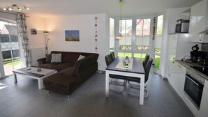 A modern living room with a sofa, coffee table, and dining table. There is a kitchen area with appliances and a view of the outside. - Erdgeschosswohnung "godewind" Zentral in Greetsiel (Greetsiel)