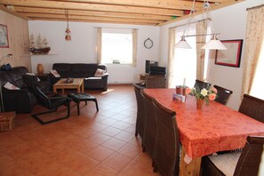Ein gemütliches Wohnzimmer mit einer Couch, Sesseln, einem Esstisch mit roter Tischdecke, einer Schiffdekoration und einer Wanduhr.