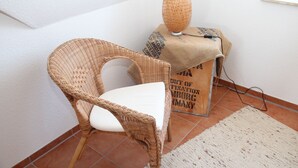 Une chaise en osier avec un coussin blanc, une lampe en fibres tressées et un sac en toile de jute orné de texte, le tout posé sur un sol carrelé.