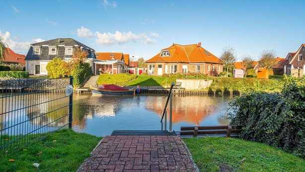 A canal with a boat docked on the left, surrounded by houses with red roofs and a grassy area in the foreground. - Schöne Ferienwohnung in Wassernähe (Greetsiel)