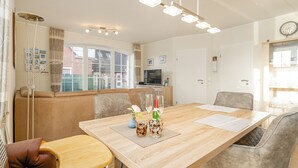 A modern living room with a long wooden table, comfortable seating, a TV, and a large window with a view of a brick building. - Schöne Ferienwohnung in Wassernähe (Greetsiel)