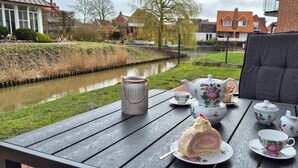 Een gedekte tafel met theeservies en taart, met uitzicht op een gracht met huizen op de achtergrond.