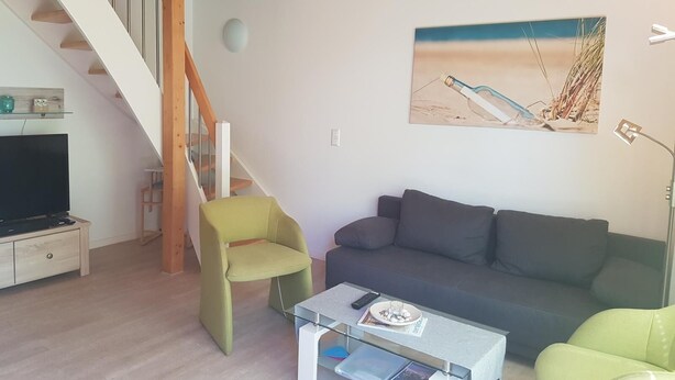 A living room with a wooden staircase, a grey sofa, a green armchair, and a painting of a beach scene.