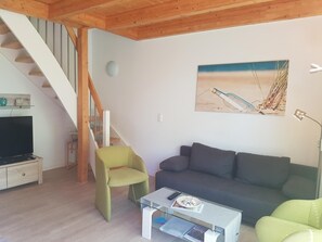 Un salon avec un escalier en bois, un canapé gris, un fauteuil vert et un tableau représentant une scène de plage.