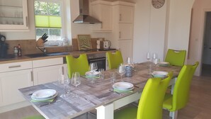 A dining area with a wooden table set for six, surrounded by green chairs. There is a window with blinds, a kitchen area with white cabinets, and a clock on the wall.
