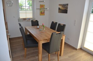 Uma sala de jantar com mesa de madeira e seis cadeiras, um relógio na parede e uma janela com persianas.