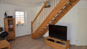 A living room with a wooden staircase, a television on a wooden stand, a bookshelf, and a window with blinds. - Allergikerfreundliches Ferienhaus in Hilgenriedersiel (Hilgenriedersiel-Hagermarsch)