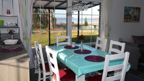 A dining area with a table set for four, white chairs, a teal tablecloth, and a view of a field through large windows. - Ferienhaus mit Traumhaftem Seeblick (Warin)