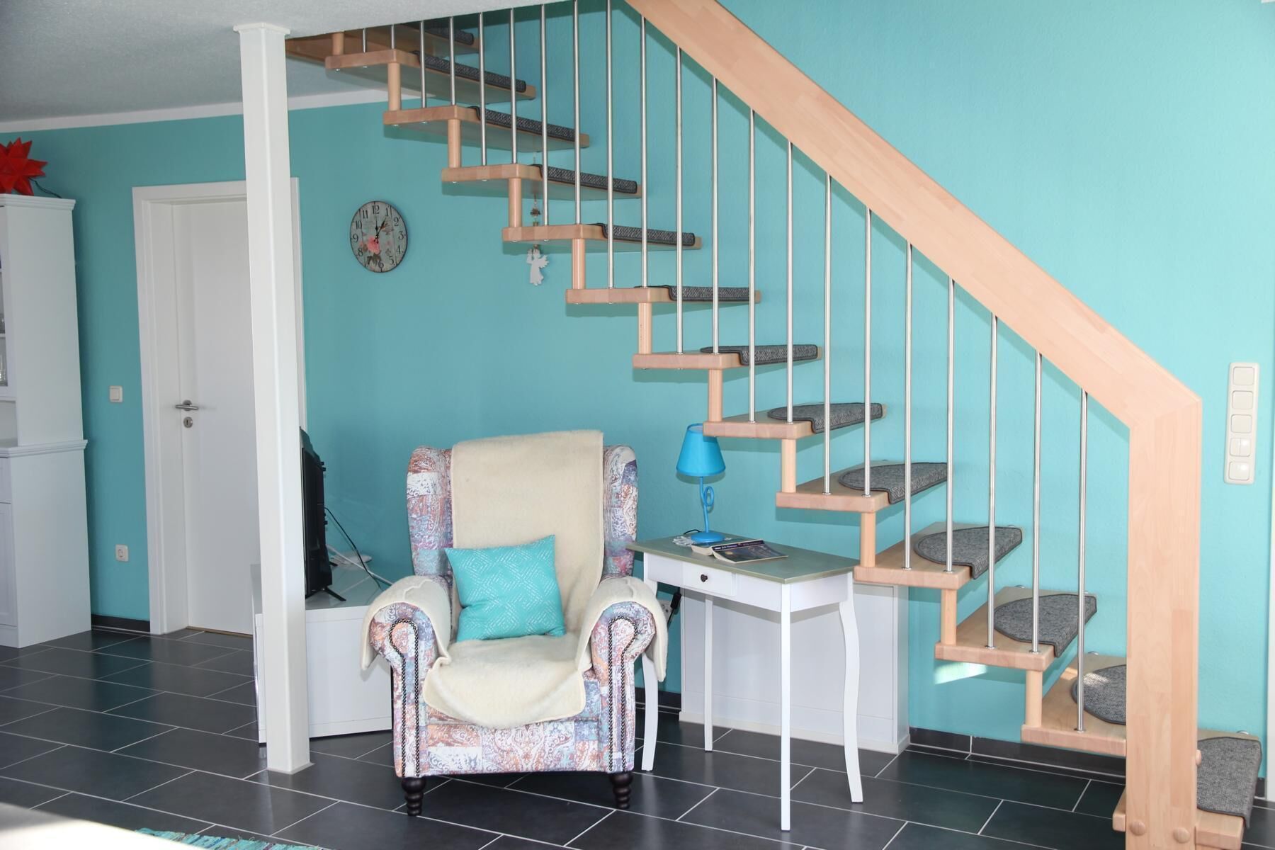 A living room with a patterned armchair, a small table with a lamp, and a wooden staircase against a teal wall. - Ferienhaus mit Traumhaftem Seeblick (Warin)