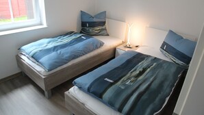 Two single beds with matching blue bedding, a bedside table with a lamp, and a window with a view of a red building.