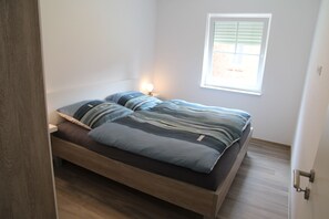 Una cama individual con estructura y cabecera de madera, una mesita de noche con lámpara, y una ventana con persianas.