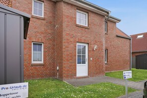 Une maison en briques rouges avec une porte et des fenêtres blanches, un petit jardin devant la maison avec de la pelouse et des fleurs, et un panneau indiquant la disponibilité de places de stationnement.