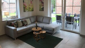 En moderne stue med en grå modulsofa, to runde træsideborde og en grøn måtte. Der er tre rammede blomstermalerier på væggen, og et vindue med et skilt, der viser 'TRAM'.
