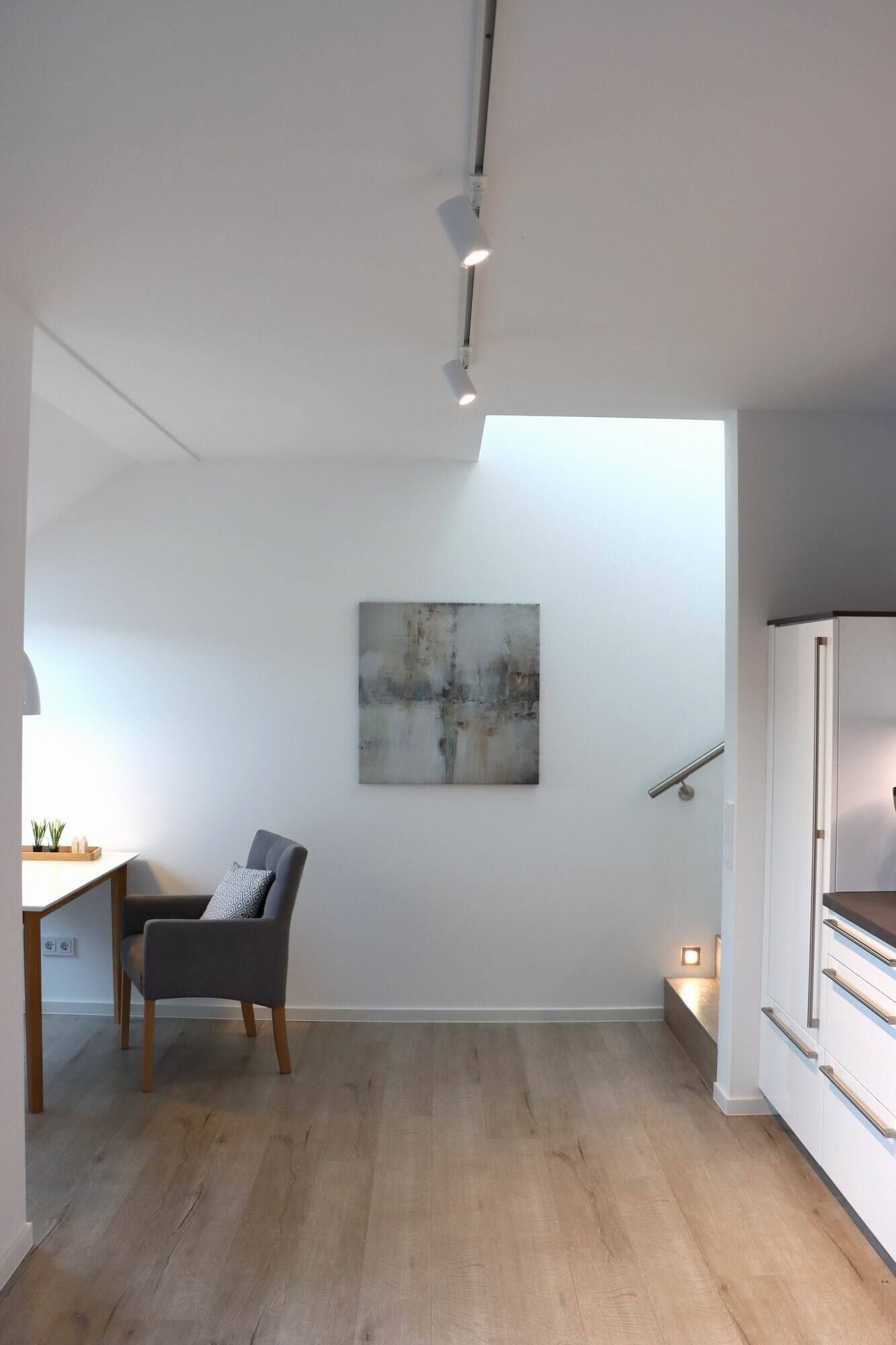 A minimalist room with a wooden floor, a grey chair, a white cabinet, and a painting on the wall.