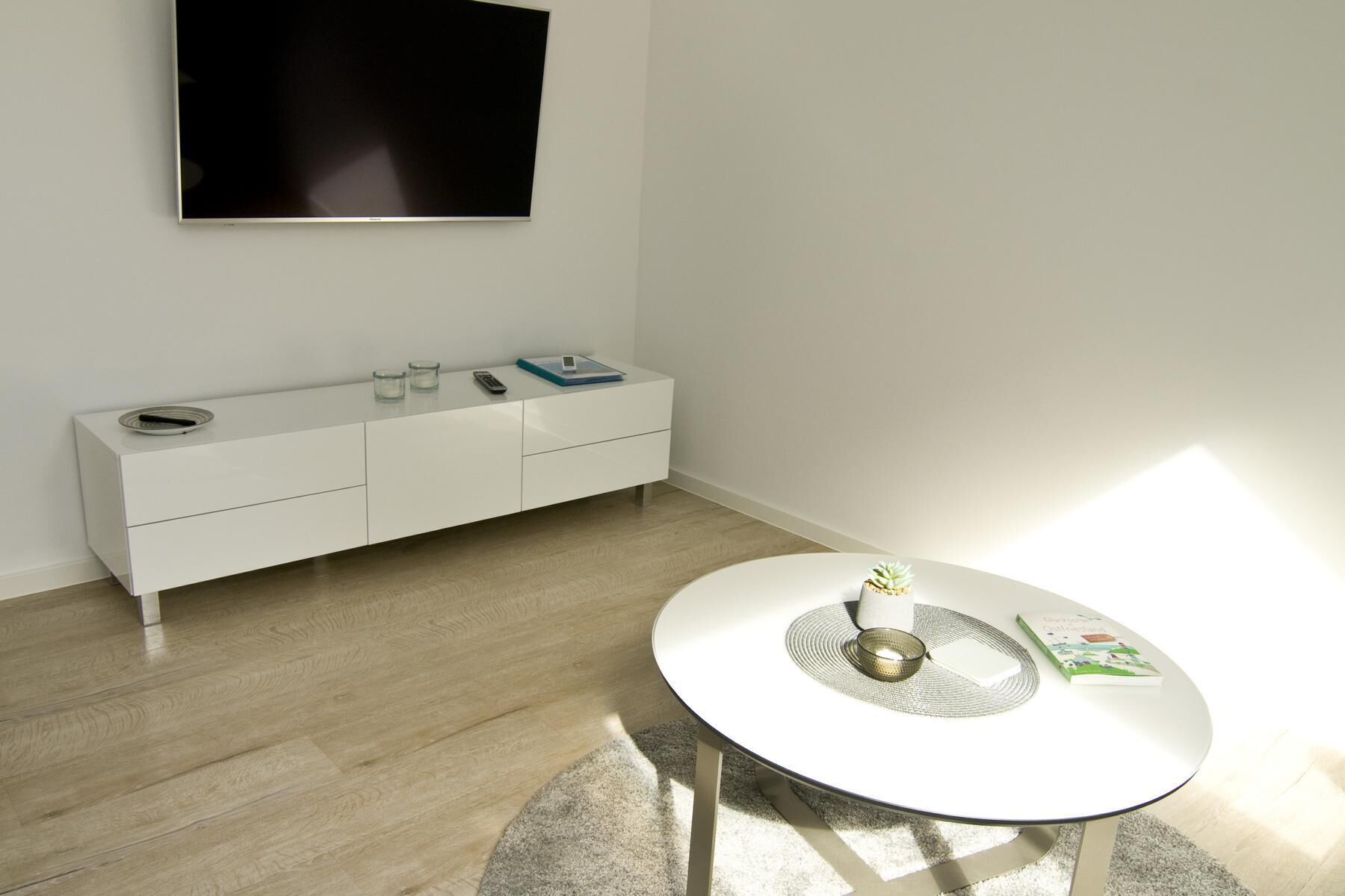 Een moderne, minimalistische kamer met een witte console tafel, een ronde tafel met een geweven placemat en een grote flatscreen televisie aan de muur.