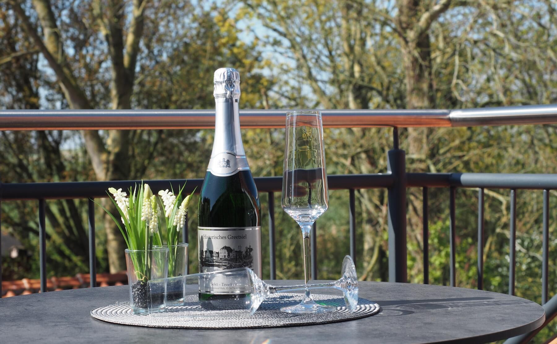 A bottle of wine, two glasses, and a small vase with flowers on a table outdoors.