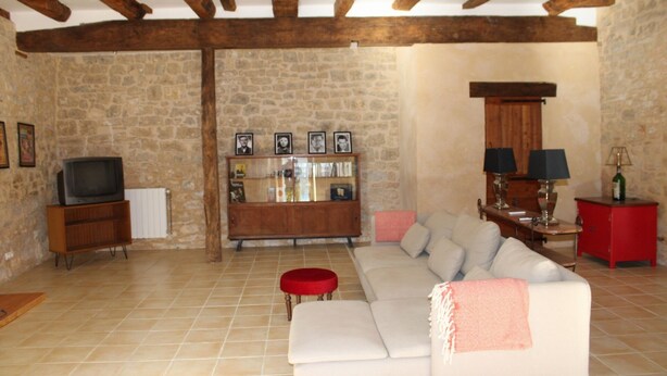 A living room with a stone wall, wooden beams, a white sofa, a red ottoman, a television, a cabinet with framed photos, and a red cabinet with lamps.