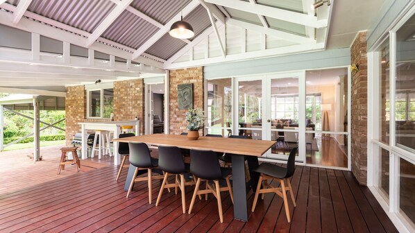 An open-plan living area with a wooden deck, a brick fireplace, and a large dining table with chairs. - Inglewood Gardens in Rosebud (Rosebud)
