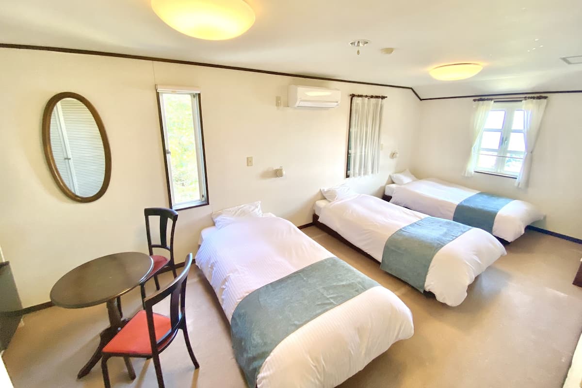 Habitación de hotel con tres camas individuales, una mesita con una silla, un espejo redondo y dos ventanas con cortinas.