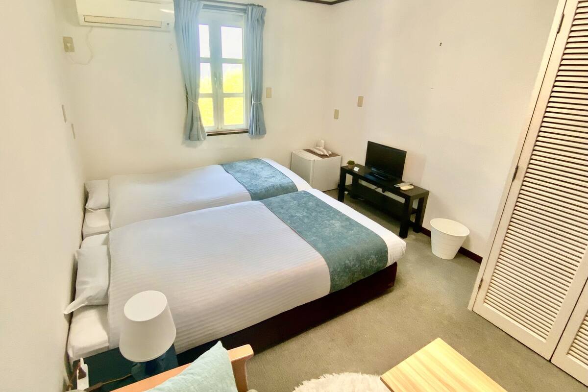 Una habitación de hotel con dos camas, televisión, una mesita y una ventana con cortinas.