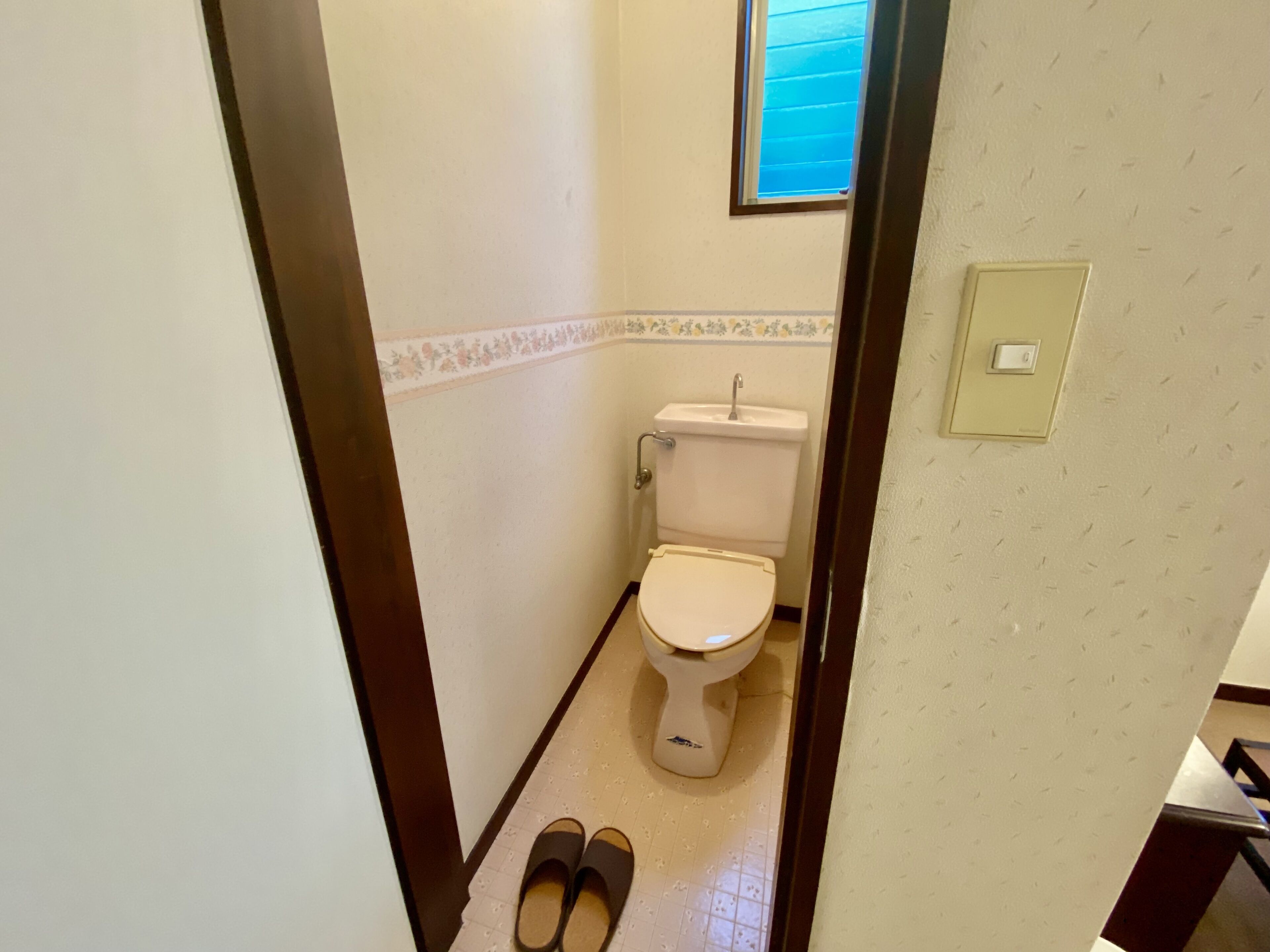 A small bathroom with a toilet, a window, and a pair of slippers on the floor.