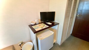 A compact room with a small desk, a television, a refrigerator, and a white bucket.