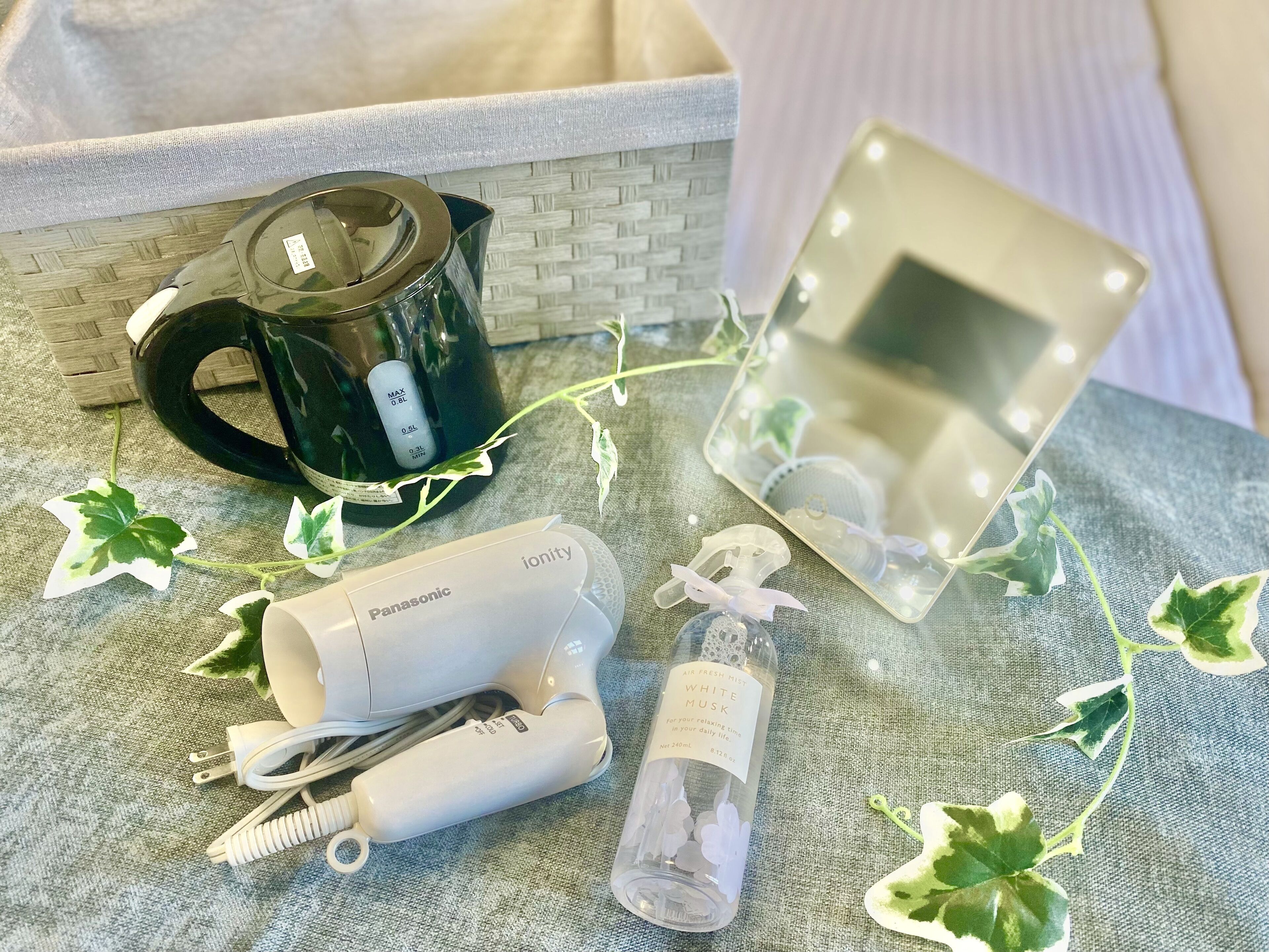 A bathroom vanity with a black kettle, a white Panasonic hairdryer, a bottle of white musk air freshener, and a mirror with lights.