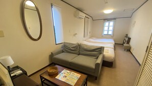 A hotel room with a sofa, a small table with a brochure, a bed, a window, and a mirror.