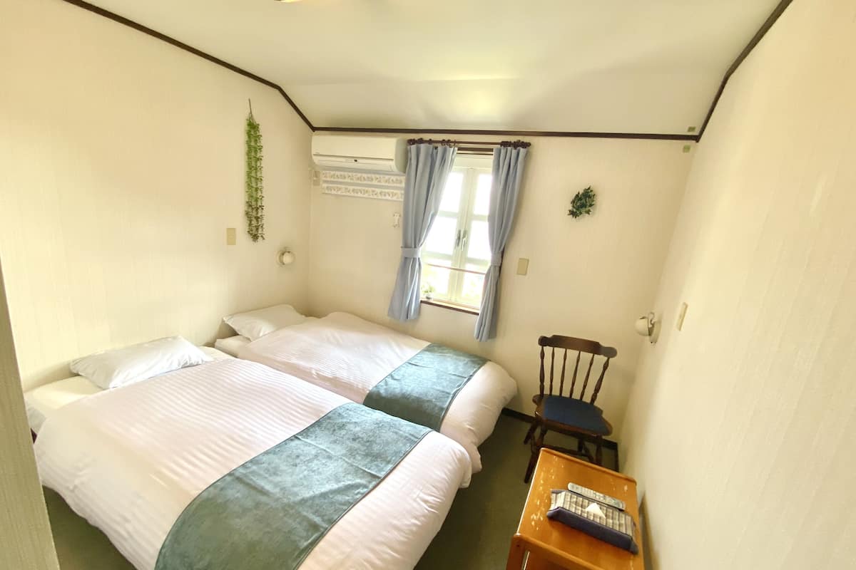 Una habitación con dos camas, una silla de madera pequeña, una mesa con un control remoto y un ventanal con cortinas.