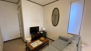 A compact living room with a sofa, a coffee table with magazines, a television on a stand, a mirror, and a window with blinds.