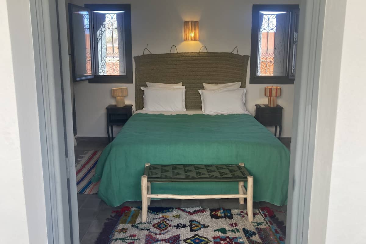 A bedroom with a bed, two bedside tables, a bench, a rug, and two windows with decorative grills.