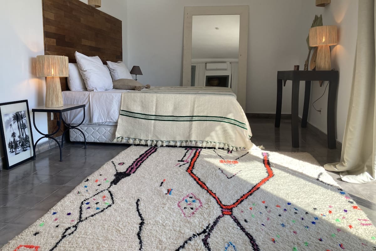 A bedroom with a wooden headboard, a bed with white bedding, a bedside table with a lamp, a framed picture, and a colorful rug.