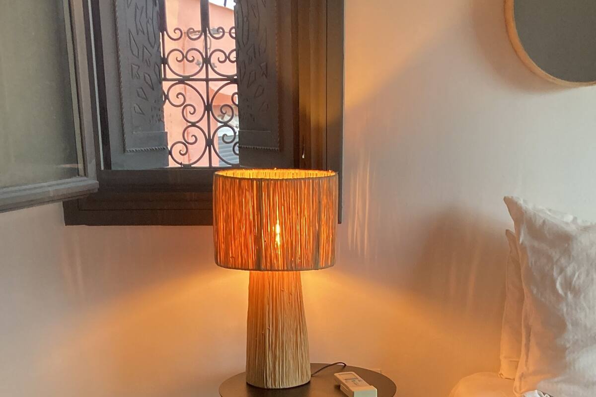 A room with a bed, a bedside table with a lamp, a patterned rug, and a window with a decorative iron grille.