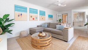 A living room with a grey sectional sofa, a round wooden coffee table, and a white brick wall with beach-themed artwork.