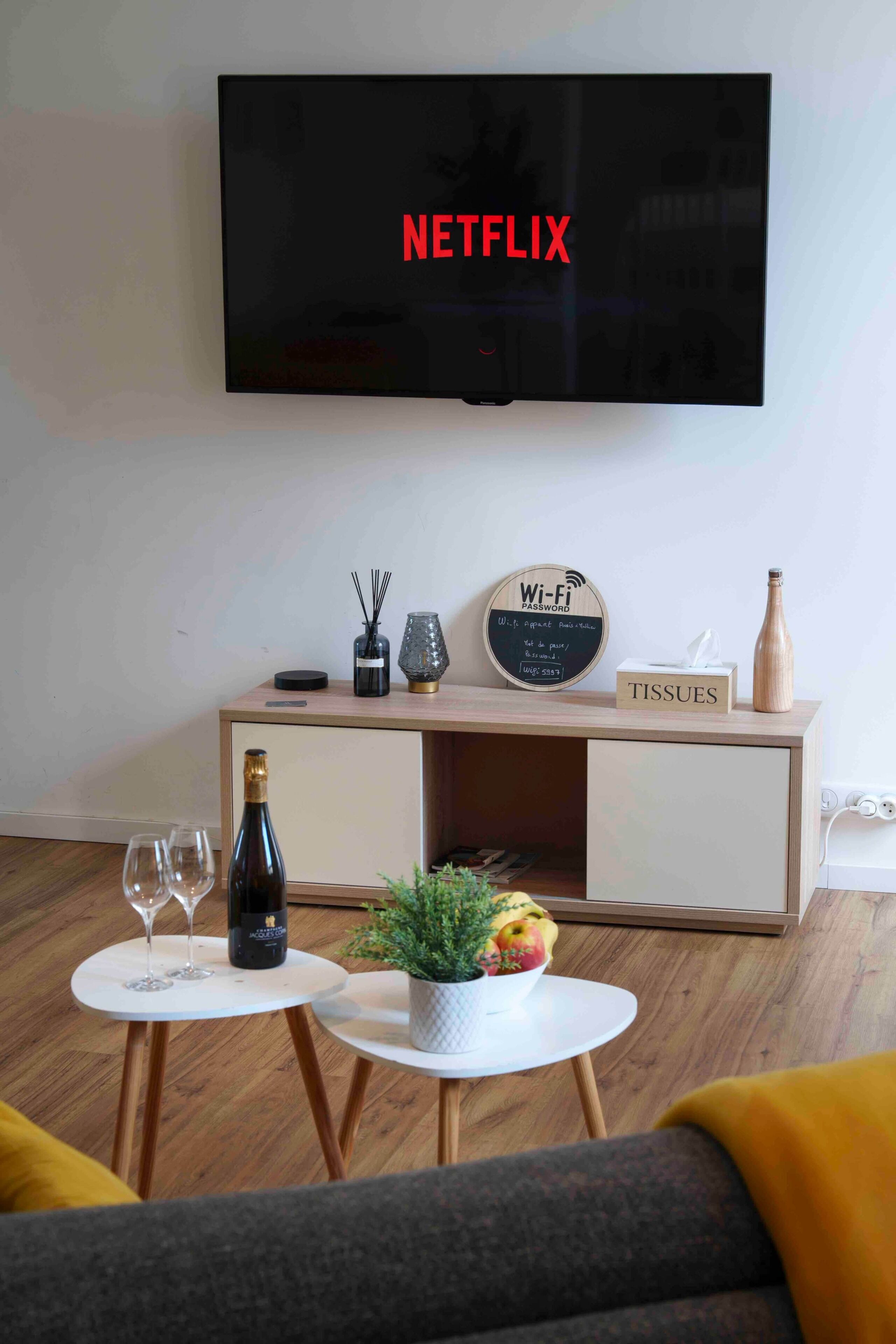 Olohuoneessa on litteänäyttöinen televisio, jossa näkyy teksti ’NETFLIX’, puinen sivupöytä, jossa on kyltti ’WI-FI-SALASANA’, paperipakkauksessa olevia paperiliinoja, pullo sekä kaksi pientä pöytää, joilla on kasvi ja hedelmiä.