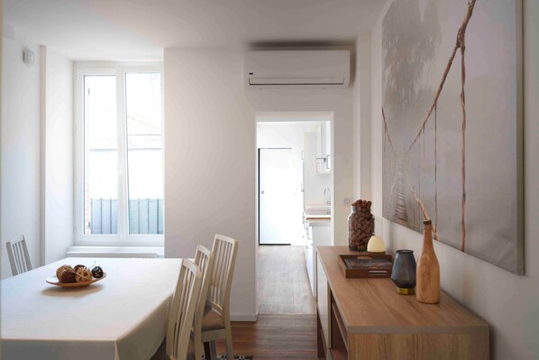 A dining area with a table set for two, a wooden side table with decorative items, and a view of the kitchen through an open door.