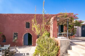 A red building with an arched doorway, a table with chairs, and a covered outdoor seating area. - The Red Winehouse | Old winery converted into a stylish villa | close to Oia (Foinikia)