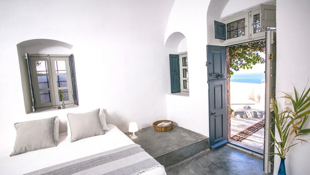 Quarto com cama, mesa de cabeceira, abajur e vista para o mar por uma porta aberta.