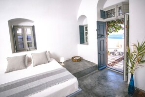 A bedroom with a bed, a nightstand, a lamp, and a view of the sea through an open door. - The Red Winehouse | Old winery converted into a stylish villa | close to Oia (Foinikia)