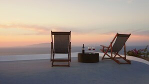Duas poltronas de madeira com almofadas, uma pequena mesa redonda com uma garrafa e dois copos, e uma vista panorâmica do oceano ao pôr do sol.