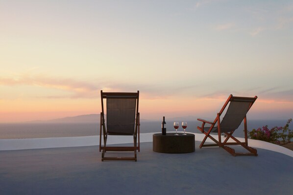 Two wooden lounge chairs with cushions, a small round table with a bottle and two glasses, and a scenic view of the ocean at sunset. - The Red Winehouse | Old winery converted into a stylish villa | close to Oia (Foinikia)