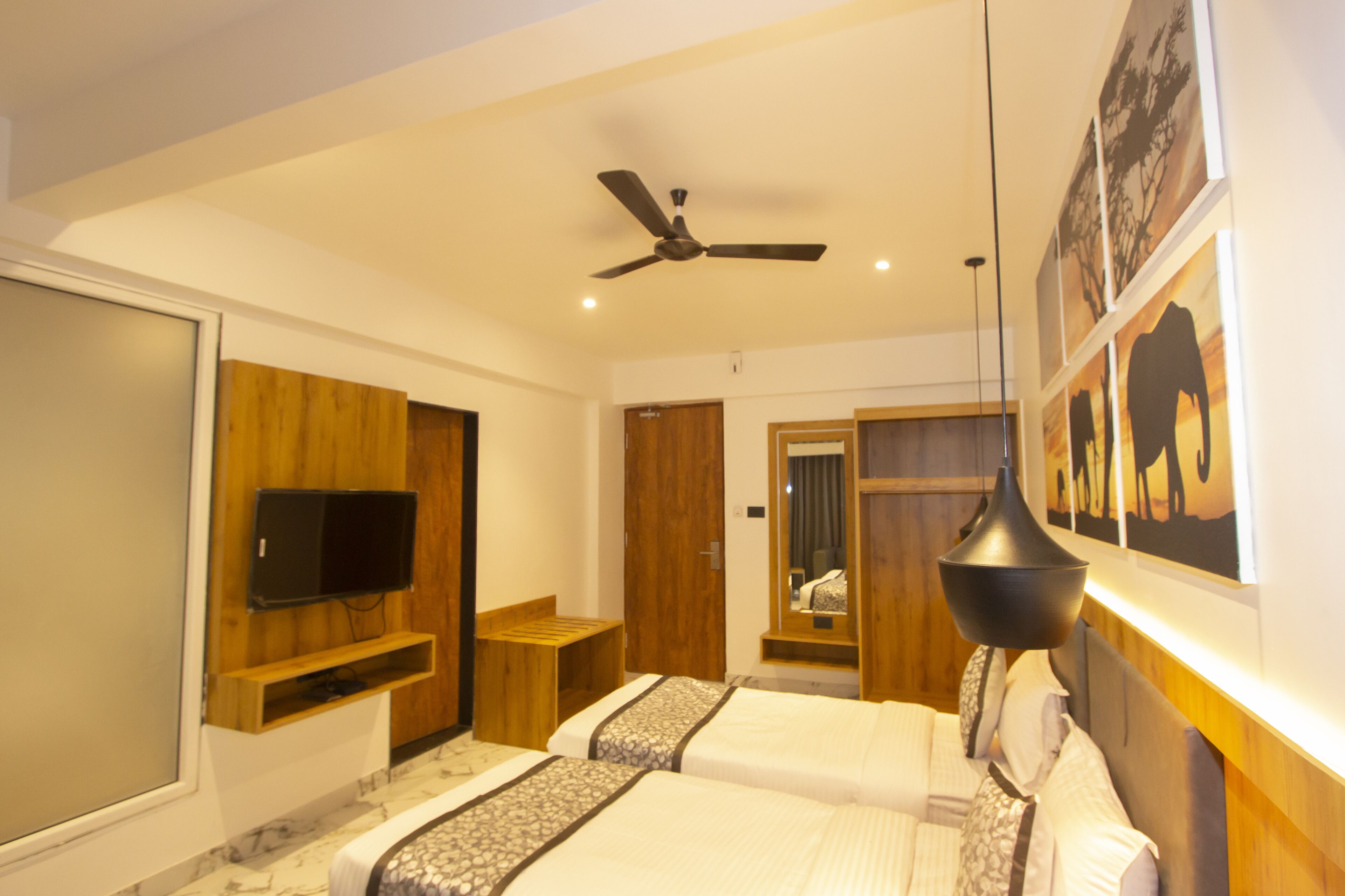 A hotel room with two beds, a ceiling fan, a mounted TV, and artwork on the walls.