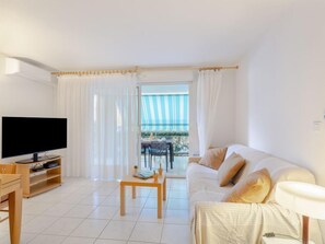 Uma sala de estar moderna com televisão de tela plana, mesa de centro de madeira, sofá branco e varanda com vista para o mar.