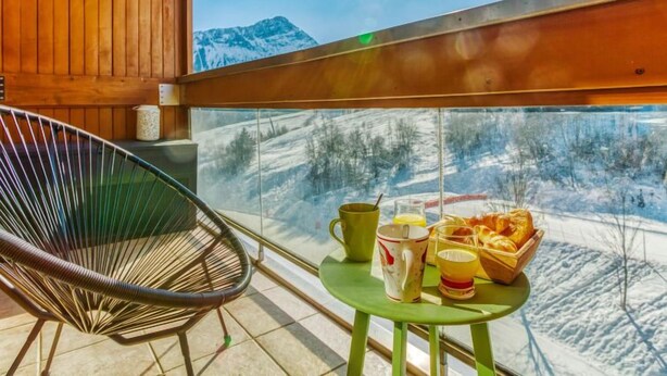 En balkong med träpanel på insidan, en stol, ett bord med frukosttillbehör och utsikt över ett snötäckt berg.