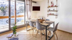 En matplats med ett bord dukat för fyra personer, ett stort fönster med gardiner, en väggmonterad TV och en hylla med juldekorationer.