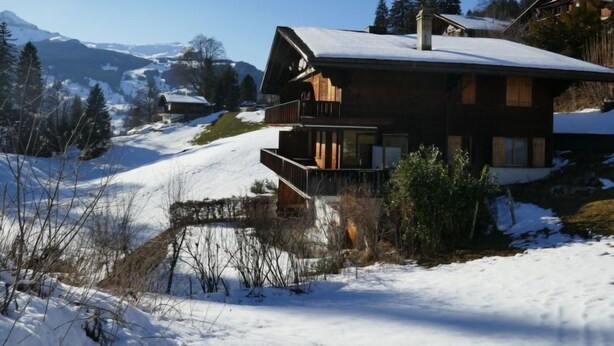 Een houten chalet met balkon, omgeven door een besneeid landschap en bergen.