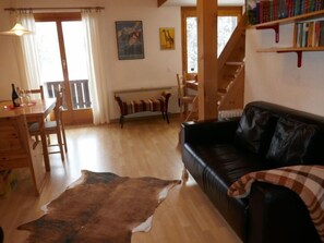 A cozy living room with a wooden dining table, a black leather sofa, a wooden bench, and a large rug.