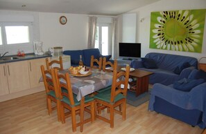A living room with a dining table, chairs, a sofa, a TV, and a kitchen area with cabinets and a window.