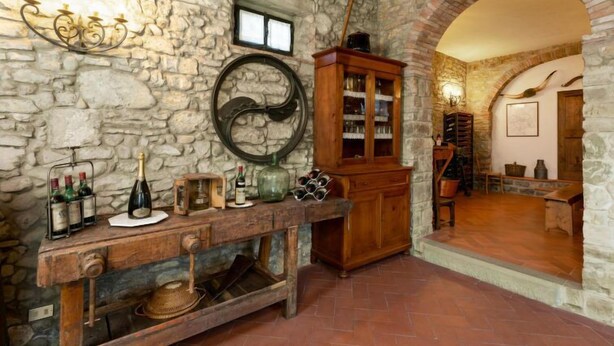 A rustic stone-walled room with a wooden table displaying wine bottles and glasses, a wooden cabinet, and a stone archway leading to another room. - Casa Mario by Interhome (Greve in Chianti)